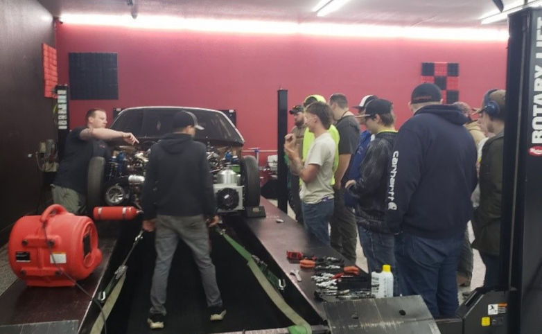 Northern Performance staff talk to students while standing around a car on a dyno in a red-walled garage, with tools and a red blower.