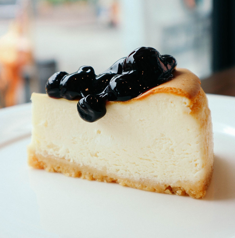 Slice of cheesecake on a white plate topped with glossy black blueberry sauce, in a soft blurred background.