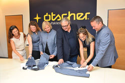 Six smiling people examine blue jackets on a table in an office, with a large yellow Ötscher logo on a navy wall.