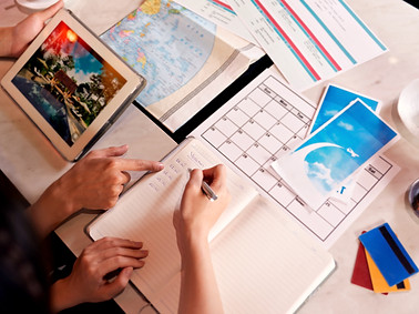 table avec une tablette, une carte du monde, un calendrier, et un cahier ouvert sur lequel écrit une personne et l'autre la conseille