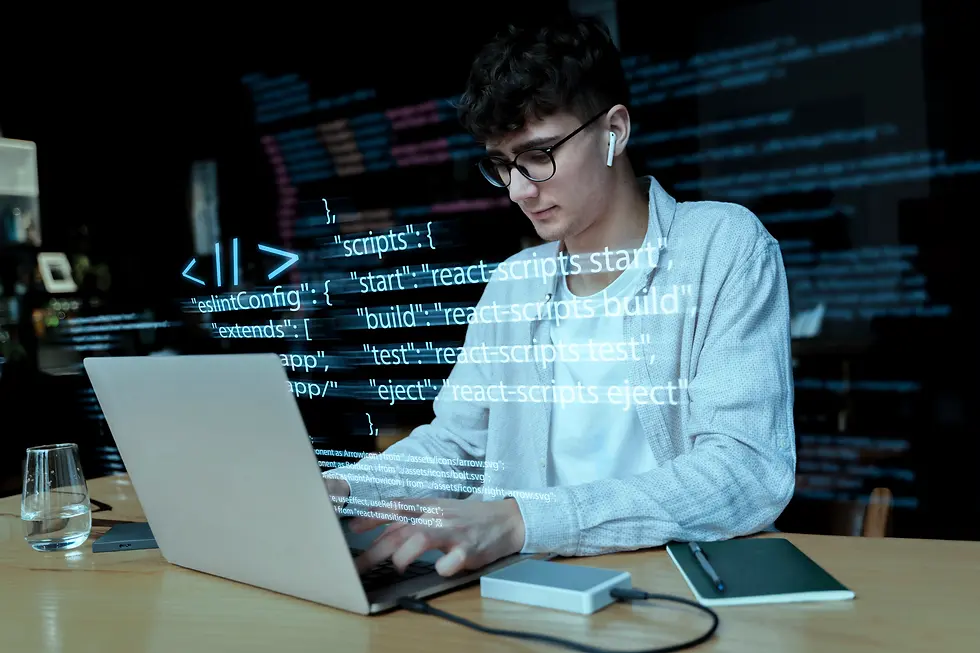 Young programmer with wireless earbuds typing code on a laptop, with a holographic projection of JavaScript commands related to React scripts displayed in front of him, in a modern office setting.