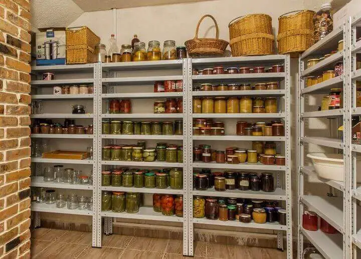 Shelves filled with colorful jars of preserved food in a pantry. Baskets and empty jars are on top. Brick wall and tiled floor visible.