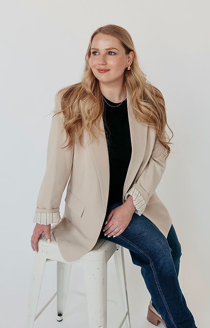 A woman in a tan blazer and black blouse sitting on a white metal stool in front of a white backdrop
