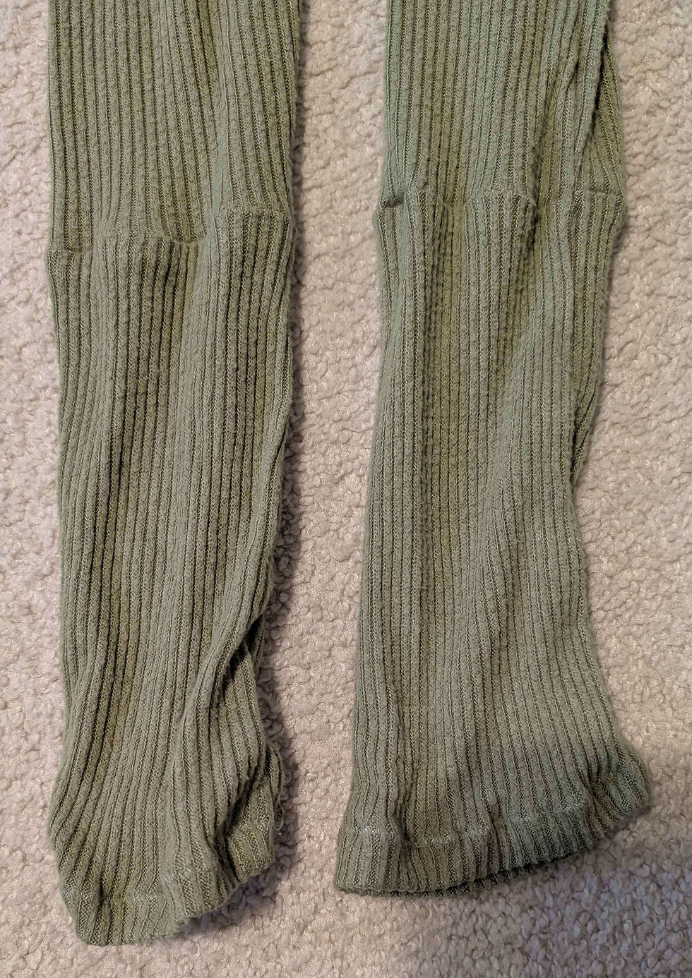Close-up of green ribbed knit pants on a beige textured surface. The fabric appears soft with a casual, relaxed look.