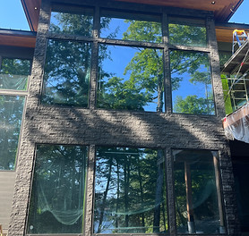 A modern two-story house with a dark stone veneer exterior and large floor-to-ceiling windows reflecting the surrounding tree