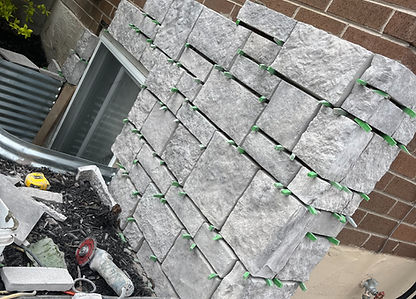 A partially completed stone veneer installation on an exterior brick wall. The light gray stone veneer pieces are arranged wi