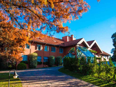 Fotografia de um casarão de arquitetura em estilo europeu e enxaimel, cercado por jardins gramados, arbustos floridos e grandes árvores sob um céu azul, representando a sede da Lapinha Clínica Spa na Lapa, Paraná.