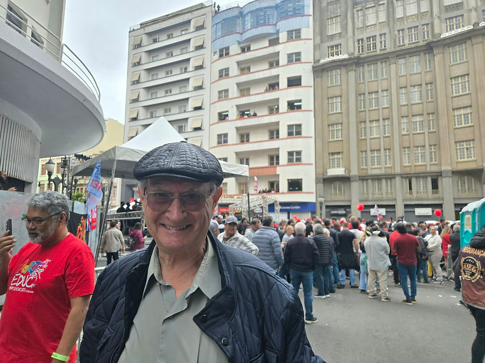 Vitório Sorotiuk sorrindo em primeiro plano, usando boina escura e jaqueta, diante de multidão reunida no calçadão da Boca Maldita em Curitiba.