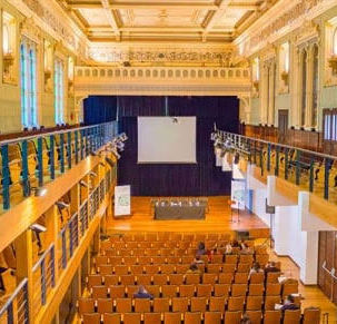 Visão interna ampla da sala de concertos da Capela Santa Maria em Curitiba. O espaço possui teto decorado com afrescos claros, galerias e camarotes laterais em madeira escura estruturados em dois andares, fileiras de poltronas estofadas voltadas para um palco ao fundo com cortinas pretas e iluminação cênica quente.
