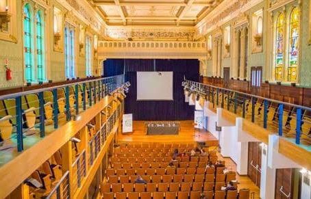 Visão interna ampla da sala de concertos da Capela Santa Maria em Curitiba. O espaço possui teto decorado com afrescos claros, galerias e camarotes laterais em madeira escura estruturados em dois andares, fileiras de poltronas estofadas voltadas para um palco ao fundo com cortinas pretas e iluminação cênica quente.