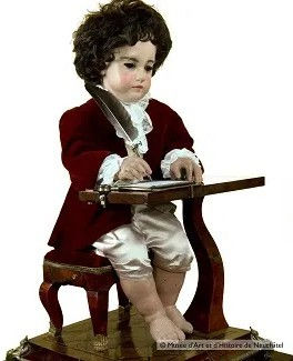 Fotografia do autômato mecânico O Escrivão do século XVIII, em formato de menino com roupas de época sentado a uma escrivaninha de madeira segurando uma pena para escrever.