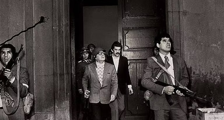 Salvador Allende vestindo capacete e segurando um fuzil enquanto olha para fora na entrada do Palácio La Moneda durante o golpe militar de 1973 no Chile.
