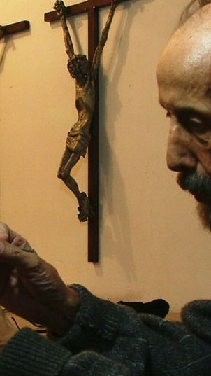 O escultor Guido Rocha em seu ateliê modelando uma peça em gesso, com três crucifixos de corpos esquálidos pendurados na parede ao fundo.