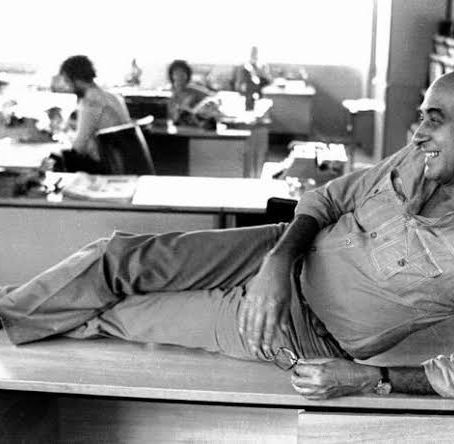 Fotografia em preto e branco do jornalista Zuenir Ventura, sorridente e deitado de lado sobre a mesa de uma redação de jornal, cercado por papéis e colegas de trabalho ao fundo.