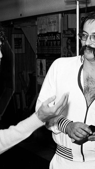 Fotografia em preto e branco de Caetano Veloso, jovem e de cabelos cacheados, gesticulando com a mão aberta enquanto conversa com Paulo Leminski, que veste agasalho esportivo claro, usa óculos e bigode marcante, em bastidores com cartaz do Projeto Pixinguinha ao fundo.