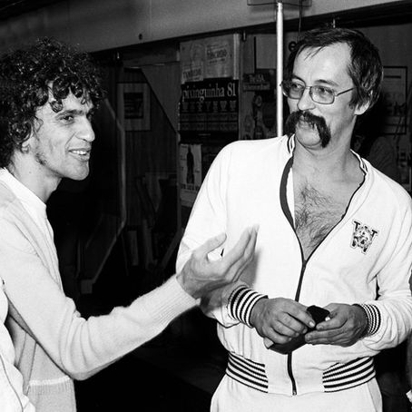 Fotografia em preto e branco de Caetano Veloso, jovem e de cabelos cacheados, gesticulando com a mão aberta enquanto conversa com Paulo Leminski, que veste agasalho esportivo claro, usa óculos e bigode marcante, em bastidores com cartaz do Projeto Pixinguinha ao fundo.