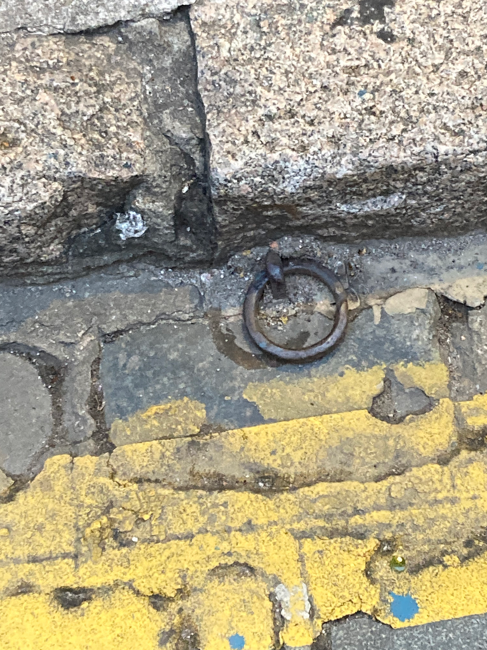 Plano detalhe de uma antiga argola de ferro rústica e oxidada cravada entre as pedras de paralelepípedo e o meio-fio com faixa amarela gasta da Rua São Francisco.