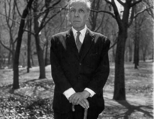 Fotografia a preto e branco do escritor Jorge Luis Borges de pé num parque com árvores despidas, vestindo fato escuro e apoiando ambas as mãos na sua bengala.