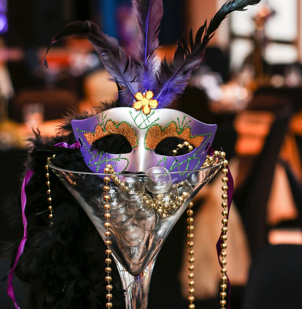 A festive purple mask with feathers and glitter sits atop a glass with beads. The background is a blurred banquet setting.