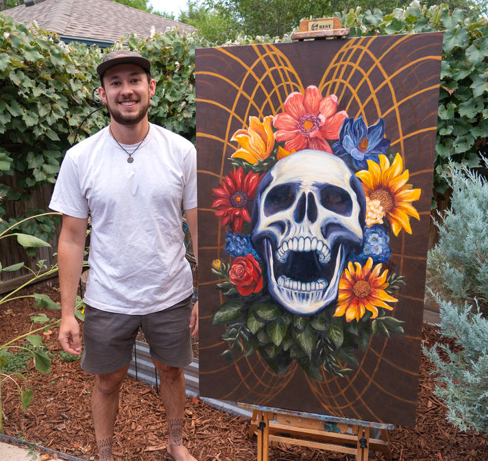 "Emergence" painting, Oil on canvas, 30"x50", featuring skull and flowers. Artist standing next to painting with a smile on their face.