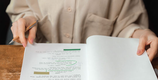 A woman wearing glasses holds a pencil while reviewing and annotating a printed script at a wooden desk.
