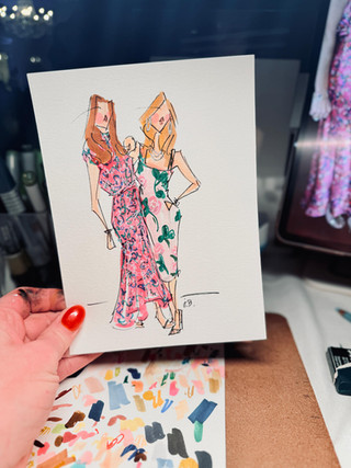 Hand holding fashion illustration of two women in floral dresses.