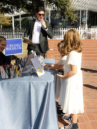 Two Young girls in white dresses interact with artist, drawing at a table, and man standing in dark suit.