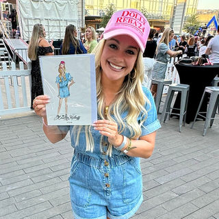 A woman in a pink hat and denim romper holds up a fashion sketch of herself, created as live art, while standing outside.