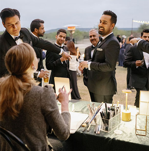 A group of men in tuxedos mingle outside where a live event artist is seated, creating wedding guest portraits.