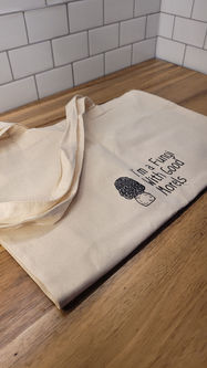 A beige canvas tote bag laying on a wood counter top. The bag has a black decal on it that says "I'm a Fungi with Good Morels" with an image of a morel mushroom