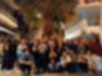 Large group of international travelers gathered under a palm tree during a hostel pub crawl in Athens, Greece. Everyone's smiling, holding drinks and capturing the social energy and connection of hostel life.