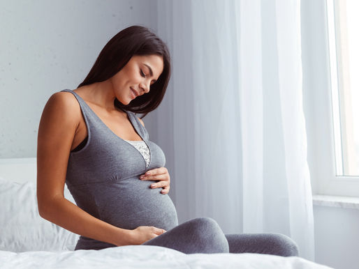 Mulher grávida sentada em uma cama perto de uma janela, vestindo uma roupa cinza, tocando suavemente a barriga e olhando para baixo com um leve sorriso.