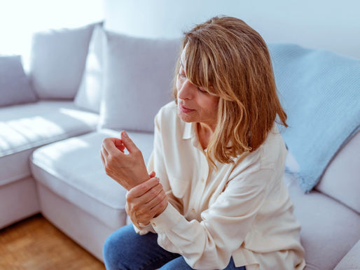 Mulher sentada no sofá segurando o punho com expressão de desconforto, representando dores articulares e saúde óssea. Ambiente claro e acolhedor, relacionado a exames de densitometria e cuidados com a saúde feminina.