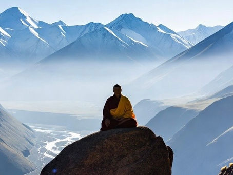 Monk in yellow and red robes meditates on a rock. Snow-capped mountains and a winding river in the background. Calm, serene setting.