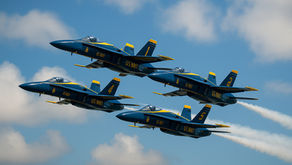 Four blue angels fighter jets, marked "U.S. Navy," performing an aerial formation against a blue sky with clouds.