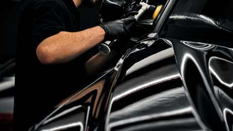 A man in black cap polishing a shiny black car with a buffer in a dimly lit garage. Reflections create dynamic patterns on the car's surface.
