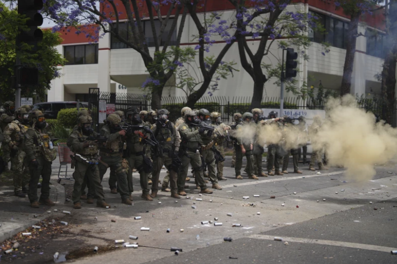 Riot police in tactical gear fire smoke grenades on a debris-littered street outside a building with trees nearby, creating a tense atmosphere.