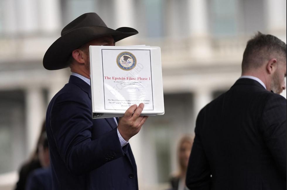 Man in suit and cowboy hat holds binder labeled "The Epstein Files: Phase 1" outside, others in background, blurred building facade.