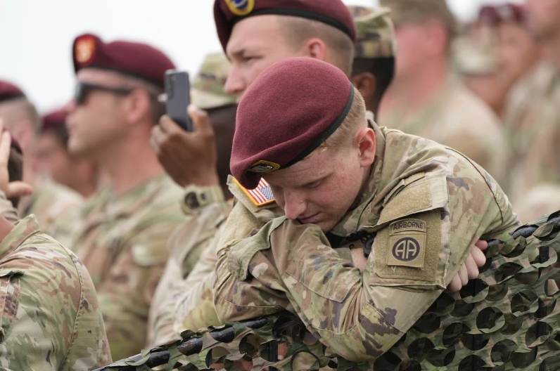 Soldiers in camouflage uniforms and maroon berets rest on a camo net. One has a patch marked Airborne. Background shows several blurred soldiers.