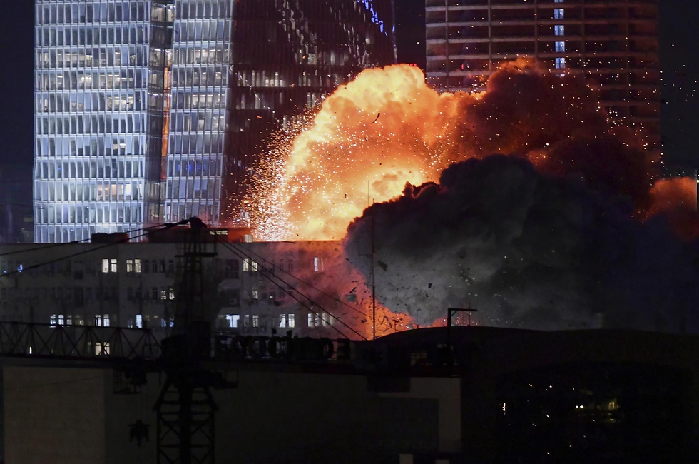 Explosion with bright orange flames and dark smoke erupts near tall reflective buildings at night, creating an intense scene.