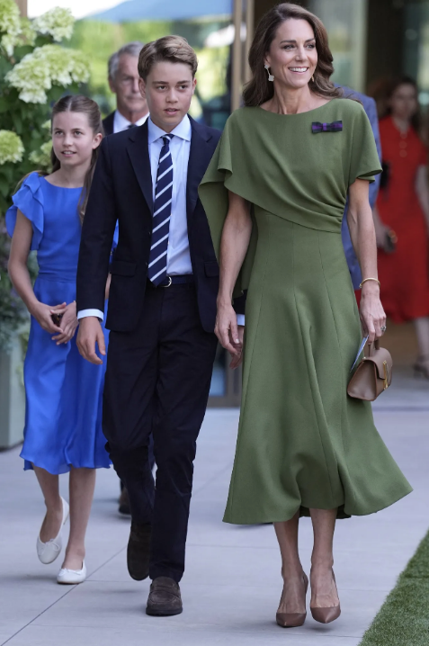 Smiling woman in green dress walks with a teen boy in a navy suit and girl in a blue dress along an outdoor path.