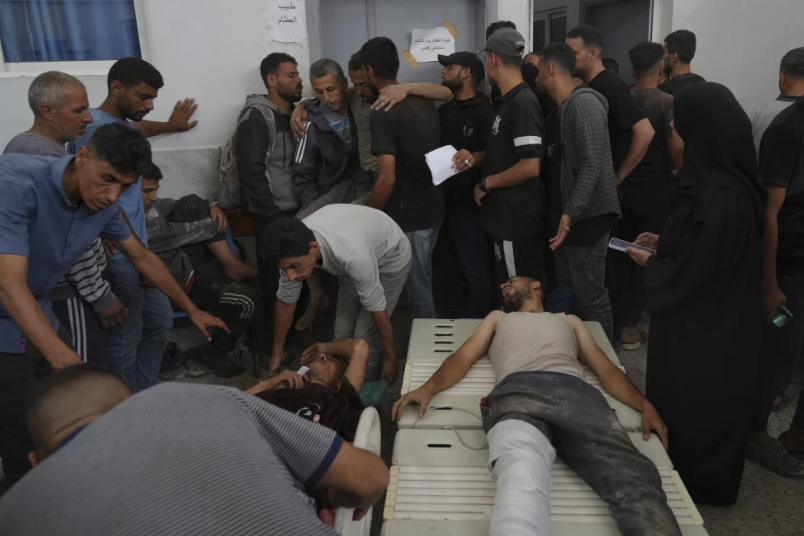 Crowded room with injured people on stretchers, others standing anxiously. Dim lighting, somber mood. Arabic text on wall background.