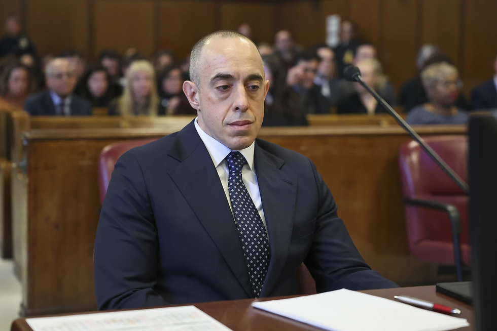 A man in a suit sits solemnly in a courtroom, surrounded by an audience. Papers are on the desk. Background is wooden and muted.