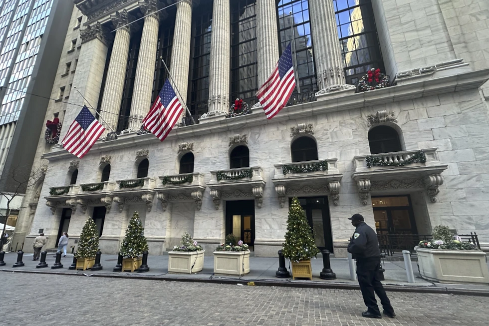 NYSE building with columns, three U.S. flags, and holiday wreaths. Decorated Christmas trees line the entrance. A security guard stands nearby.