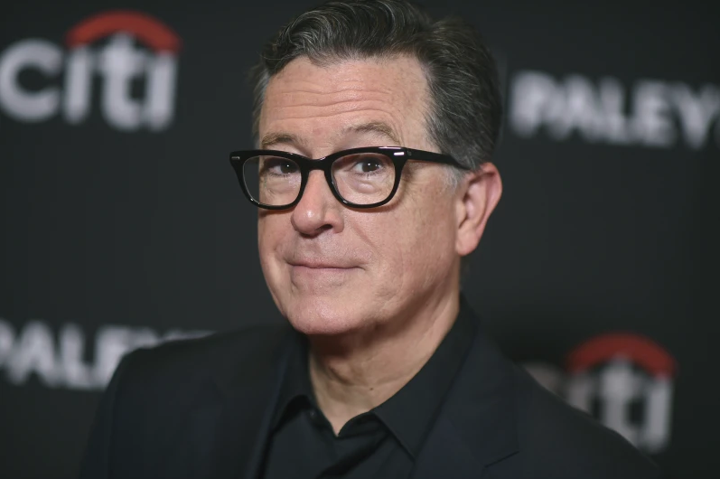 Stephen Colbert with glasses smirking, wearing a black suit. Black background with visible white text "citi" and "PALEY." Mood is confident.