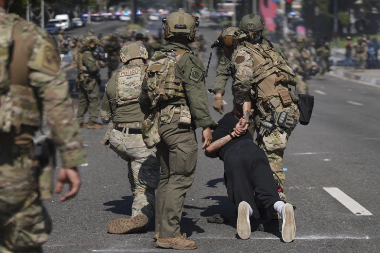Soldiers in camouflage and tactical gear detain a person on a city street. Police vehicles and more soldiers are visible in the background.