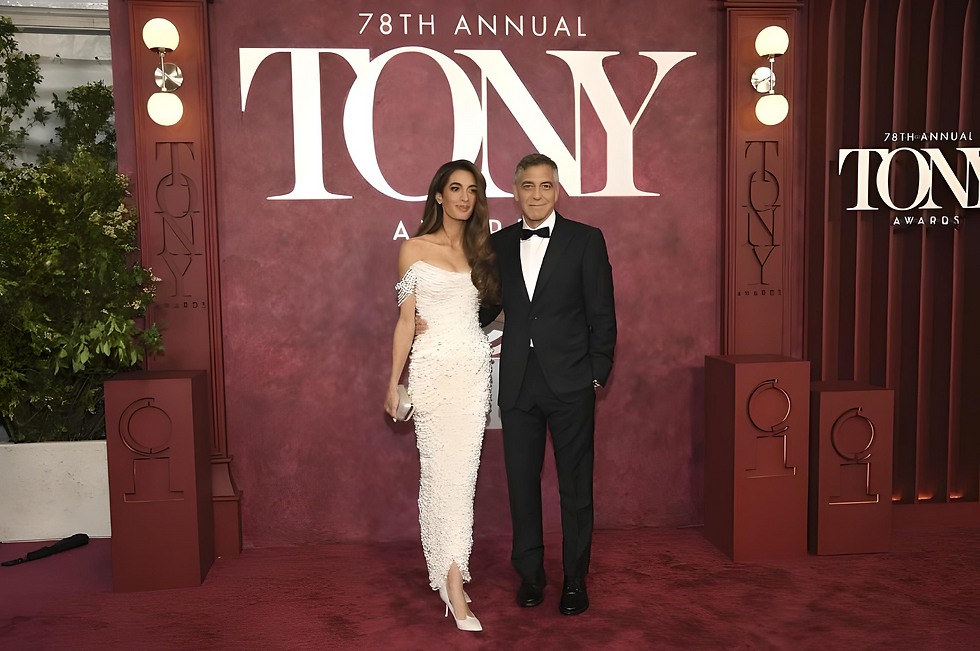 A couple poses at the 78th Annual Tony Awards in formal attire. The woman wears a white gown; the man is in a black suit. Maroon backdrop.