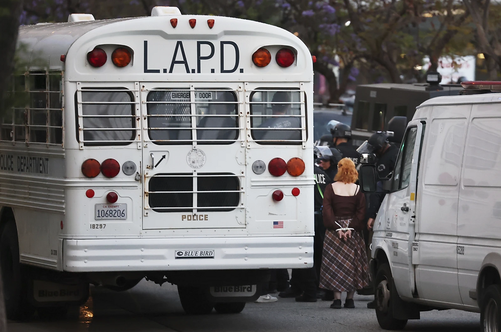 White LAPD bus on a street, officers in riot gear detaining a person with orange hair. Vans nearby, trees and buildings in the background.