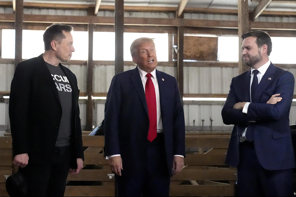 Three men stand in a wooden barn, one in a blue suit speaking, the others in casual and formal attire listening. Daylight filters in.