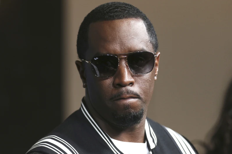 P Diddy in sunglasses and a black jacket with white stripes stands against a neutral background, displaying a serious expression.